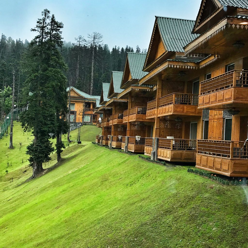 Row of multi-story wooden chalet-style buildings with peaked green roofs and balconies set on a grassy hillside, calm and inviting mood