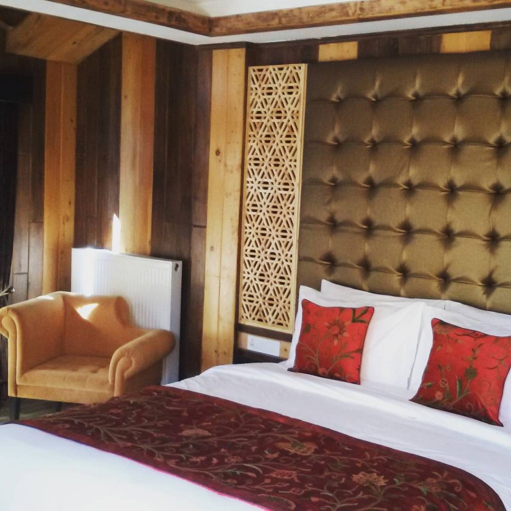 Cozy bedroom corner with a tufted headboard, red floral pillows, a mustard armchair, and wood paneled walls with a decorative window