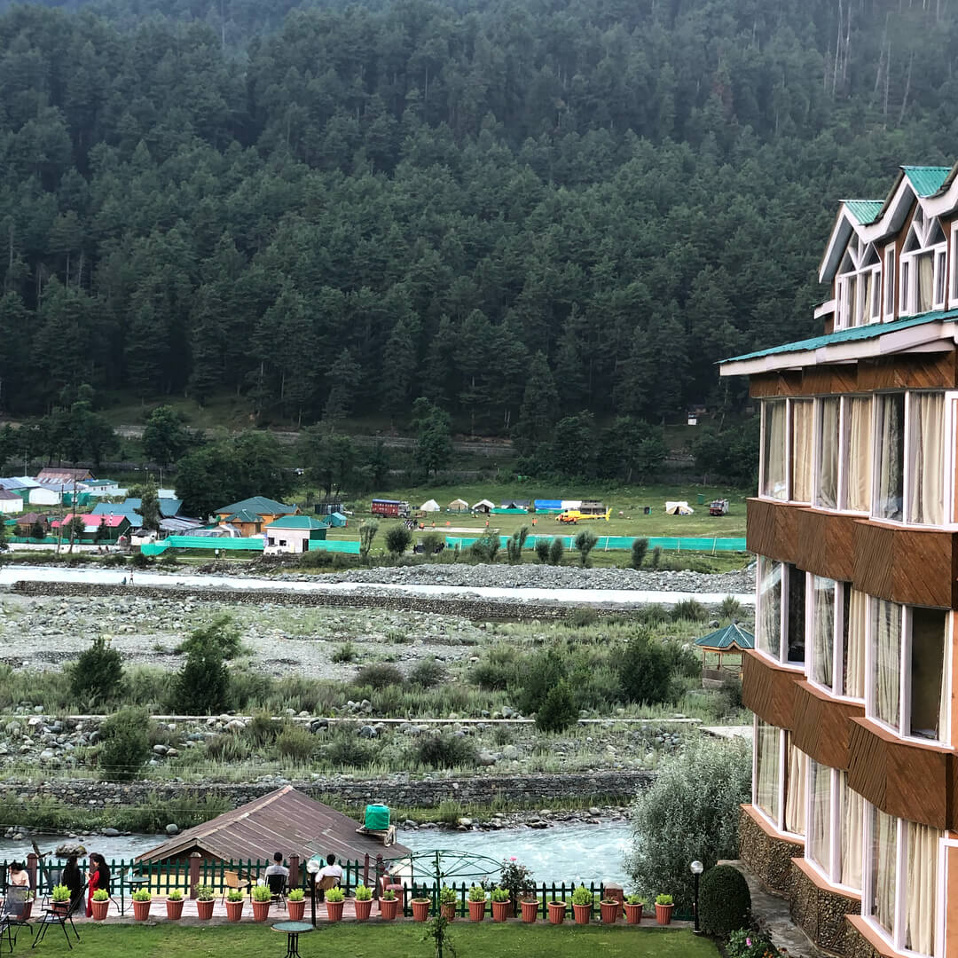 Scenic river valley with green forested hills, a multi-story building, and outdoor seating, calm and inviting mood